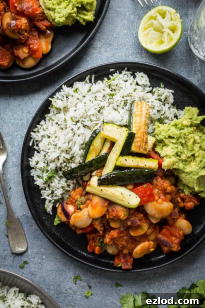 A detailed close-up of a single black plate showcasing a perfectly portioned serving of vegan lentil, vegetable, and butter bean chilli, accompanied by fragrant coriander rice, creamy guacamole, and tender roasted courgette.