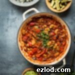 A pan of vegan vegetable, lentil and butter bean chilli with a bowl of coriander rice, roasted courgettes and guacamole on a grey background.