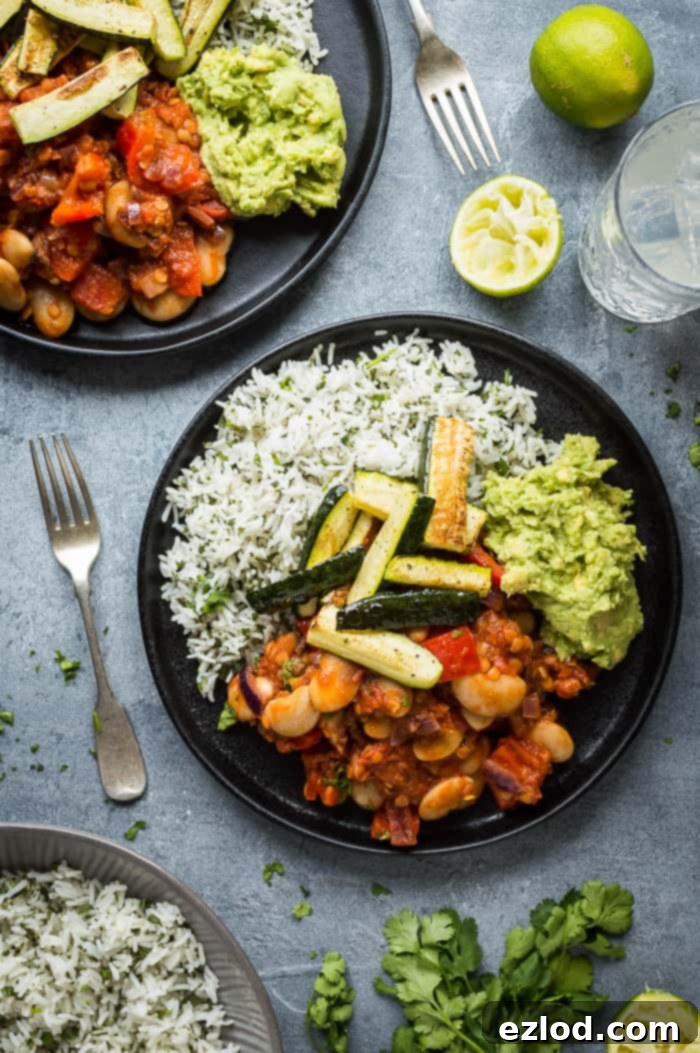 Two elegantly plated portions of vegan lentil, vegetable, and butter bean chilli, served alongside fluffy coriander rice, vibrant guacamole, and golden roasted courgette, presented on sophisticated black plates against a muted grey backdrop.