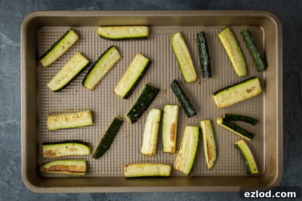 Strips of courgette (zucchini) tossed with olive oil, salt, and pepper, spread out on a baking tray, ready for roasting in the oven.