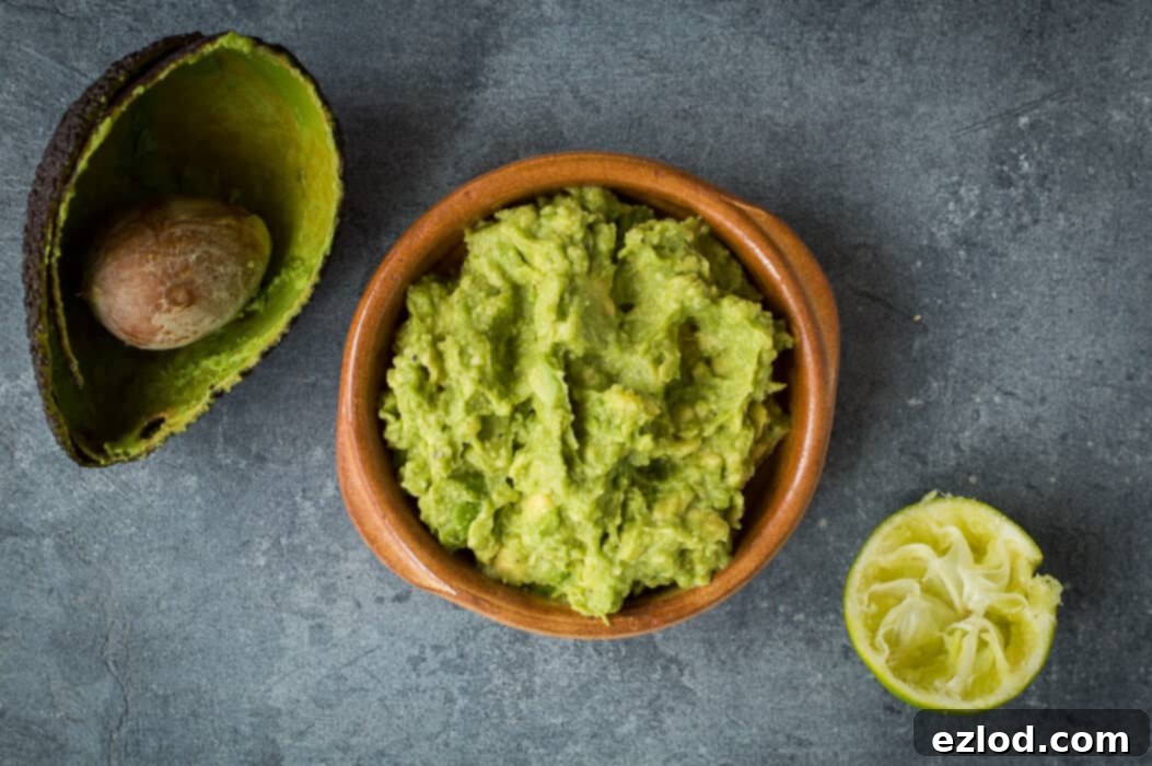 A bowl of freshly mashed avocado being prepared for guacamole, with visible lime juice, salt, and pepper being added.