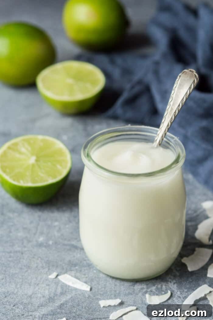 A small glass jar of homemade vegan coconut lime curd displayed on a grey background, surrounded by fresh limes and shredded coconut flakes, emphasizing its natural ingredients.