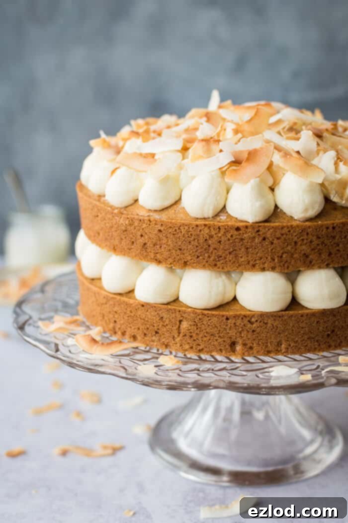 Close-up view of the side of a tall vegan coconut layer cake on a glass stand, showcasing the delightful layers of moist cake, fluffy coconut buttercream, and zesty lime curd.