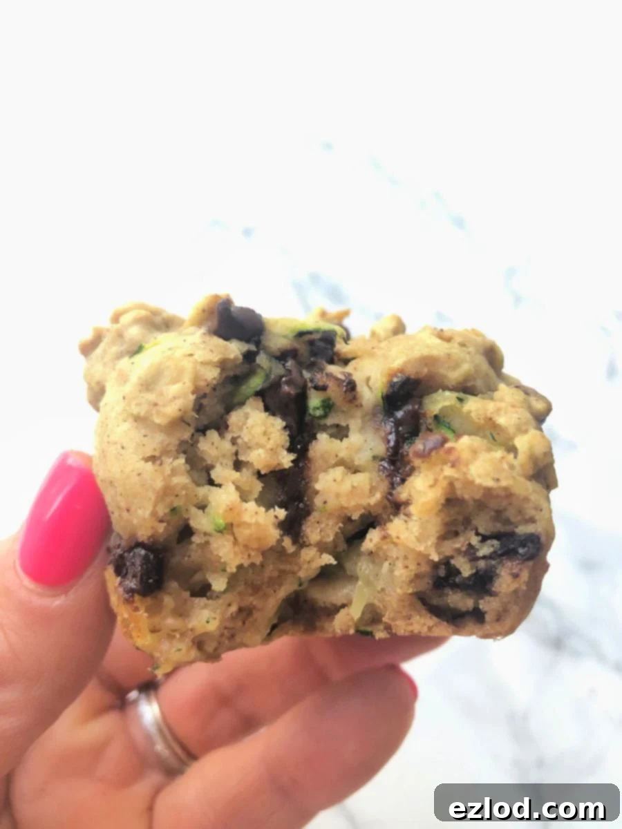 A woman's hand holding a chocolate chip zucchini muffin over a white marble background.