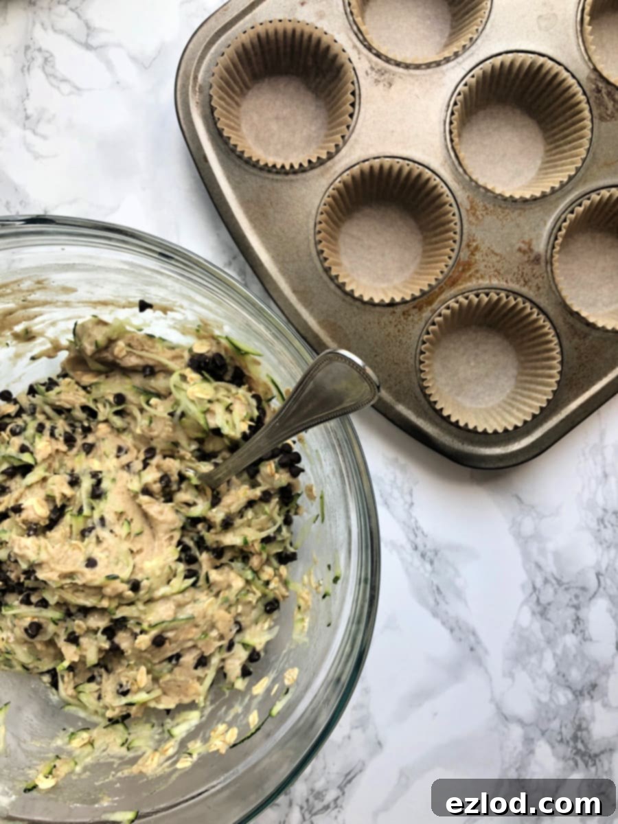 Overhead photo of Gluten free chocolate chip zucchini muffin batter in a glass bowl next to a muffin tin lined with parchment muffin liners.
