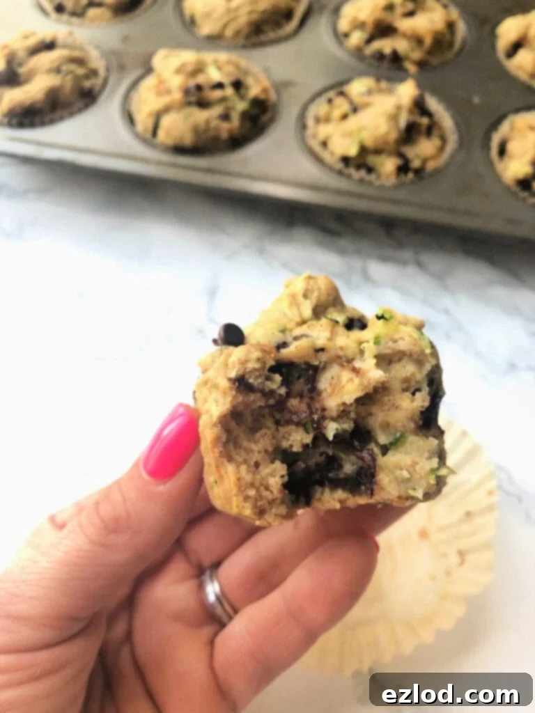 A woman's hand holding half of a zucchini muffin with the wrapper on the white counter behind it. There is also a full pan of muffins in the background slightly out of focus.