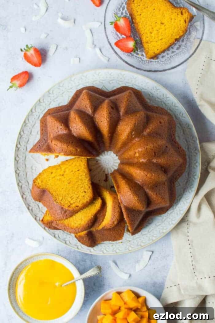 A beautifully presented vegan mango bundt cake on a white plate, set against a grey background. It's garnished with fresh strawberries, slices of ripe mango, and a small bowl of creamy mango curd, highlighting its vibrant, tropical appeal.