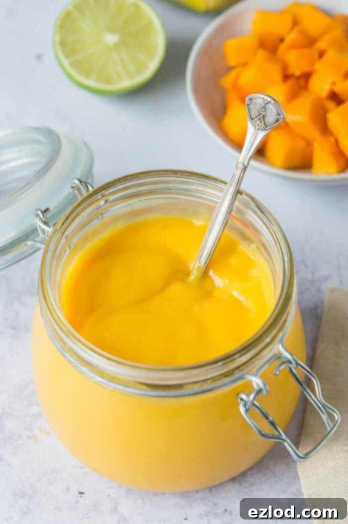 A large glass jar of eggless mango curd, beautifully presented on a grey background, accompanied by a bowl of diced fresh mango and vibrant sliced limes.