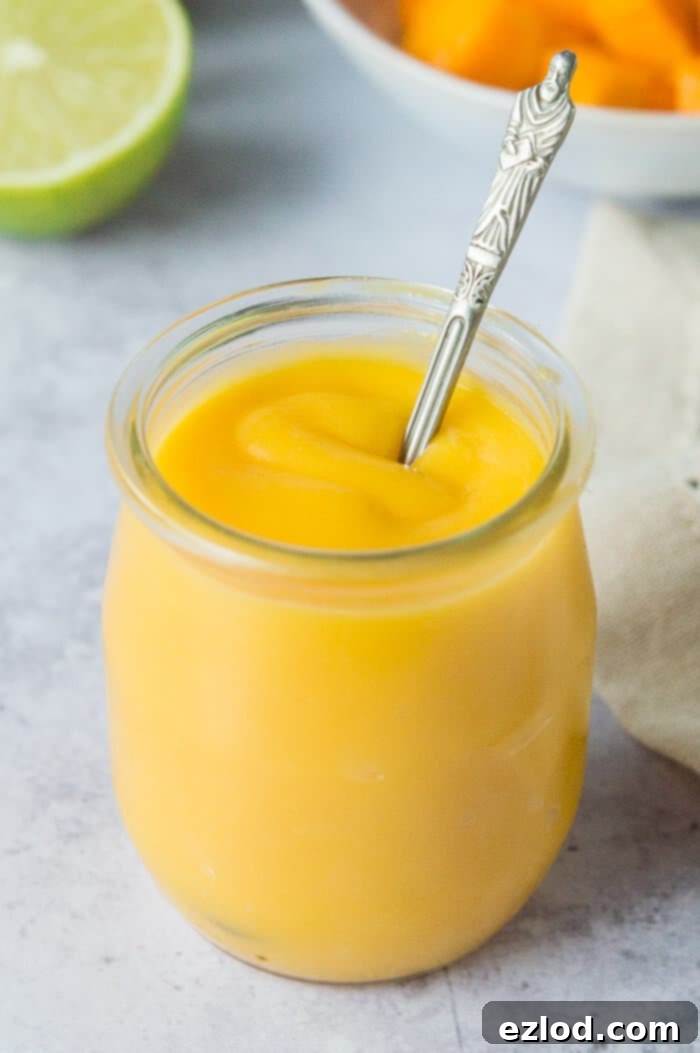 A small glass jar of vegan mango curd with a teaspoon resting inside, showcasing its rich, golden color and smooth, thick consistency.