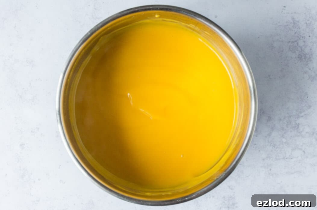 Step 2 in making vegan mango curd: The blended mixture is being cooked in a stainless steel saucepan over medium-low heat, constantly stirred with a whisk to thicken, showing its glossy surface.