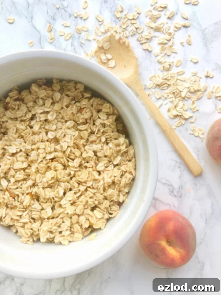 A casserole bowl of Instant Pot Peach Crisp sitting on a marble counter with a small wooden spoon and some peaches sitting nearby