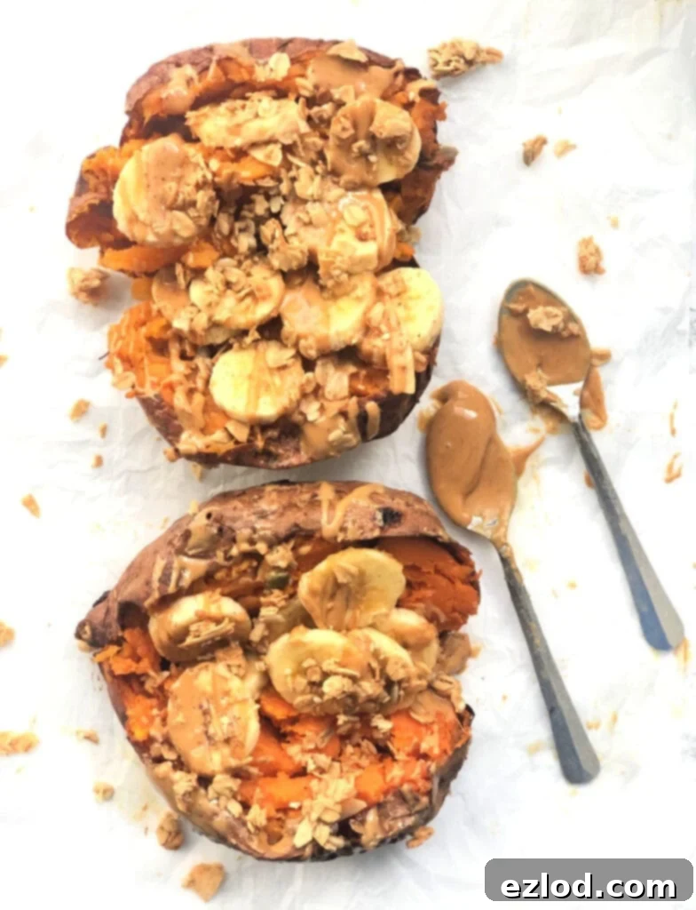 A photo of stuffed sweet potatoes on a marble background stuffed with peanut butter, bananas, and granola. Two spoons of peanut butter sit nearby