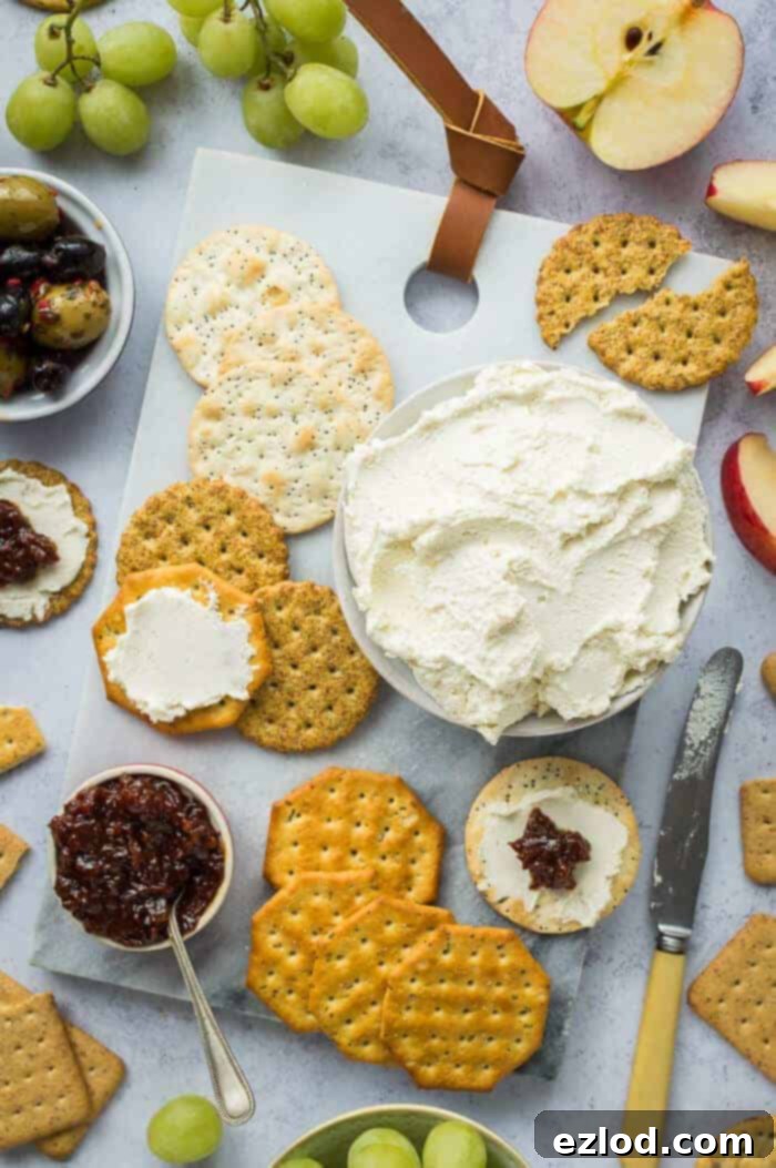 A bowl of vegan almond feta cream cheese on a marble board with crackers, chutney, olives, apple and grapes.