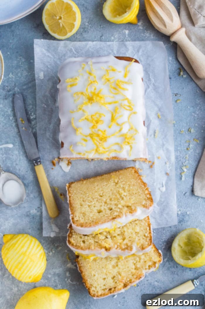 Sliced vegan lemon drizzle cake on a grey background surrounded by whole and sliced lemons, a lemon juicer, a knife and plates.