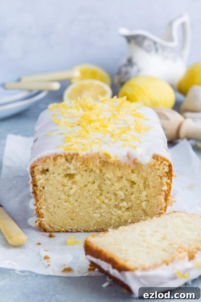 Close up of the end of a loaf of sliced lemon drizzle cake with lemons and crockery in the background.