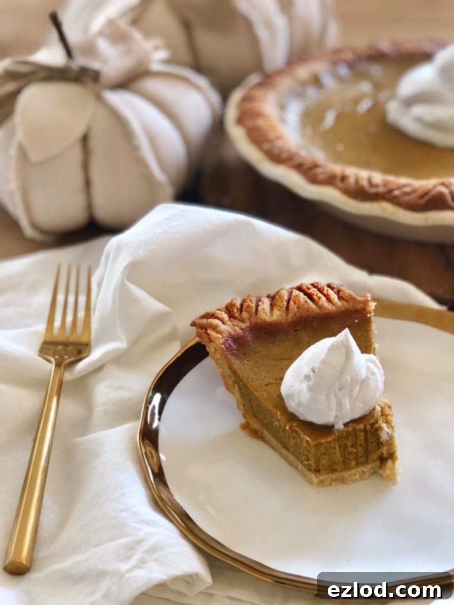 Close up photo of a partial slice of Gluten Free pumpkin pie topped with dairy-free whipped cream on a gold trimmed white plate. In the background is a whole pie and white fabric pumpkins.