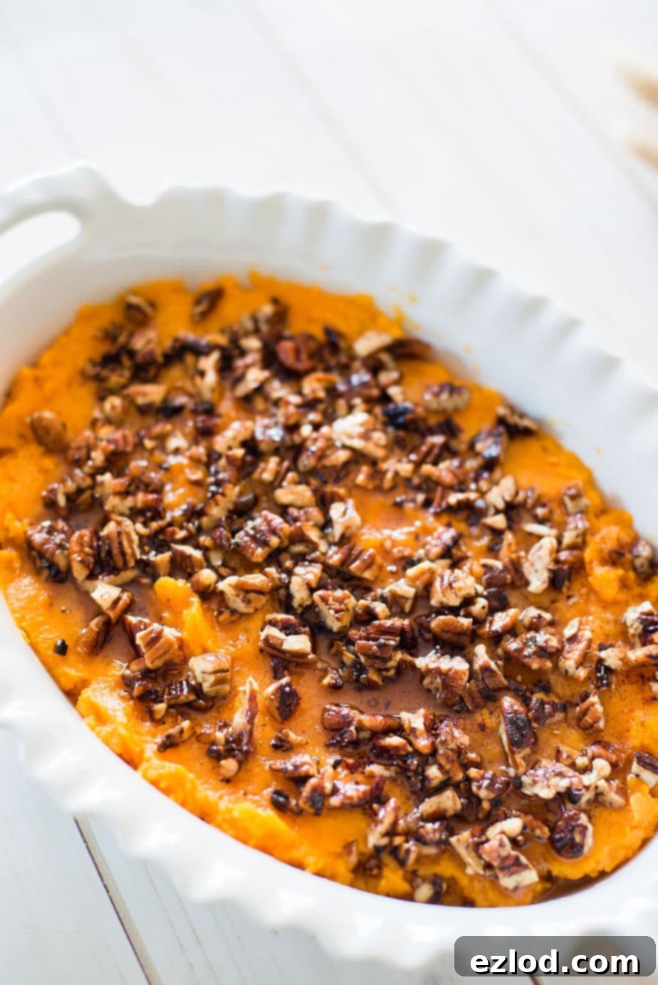 Close up photo of healthy sweet potato casserole in a white oval baking dish on a white wooden background. The mashed sweet potatoes are topped with a toasted pecan streusel.