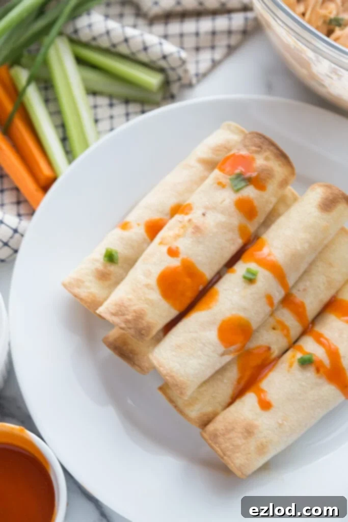 Gluten Free Buffalo Chicken Taquitos are stacked on a white dinner plate with buffalo sauce drizzled over top. In the background is a glass bowl of extra buffalo chicken filling, a ramekin of buffalo sauce, and celery and carrot sticks.