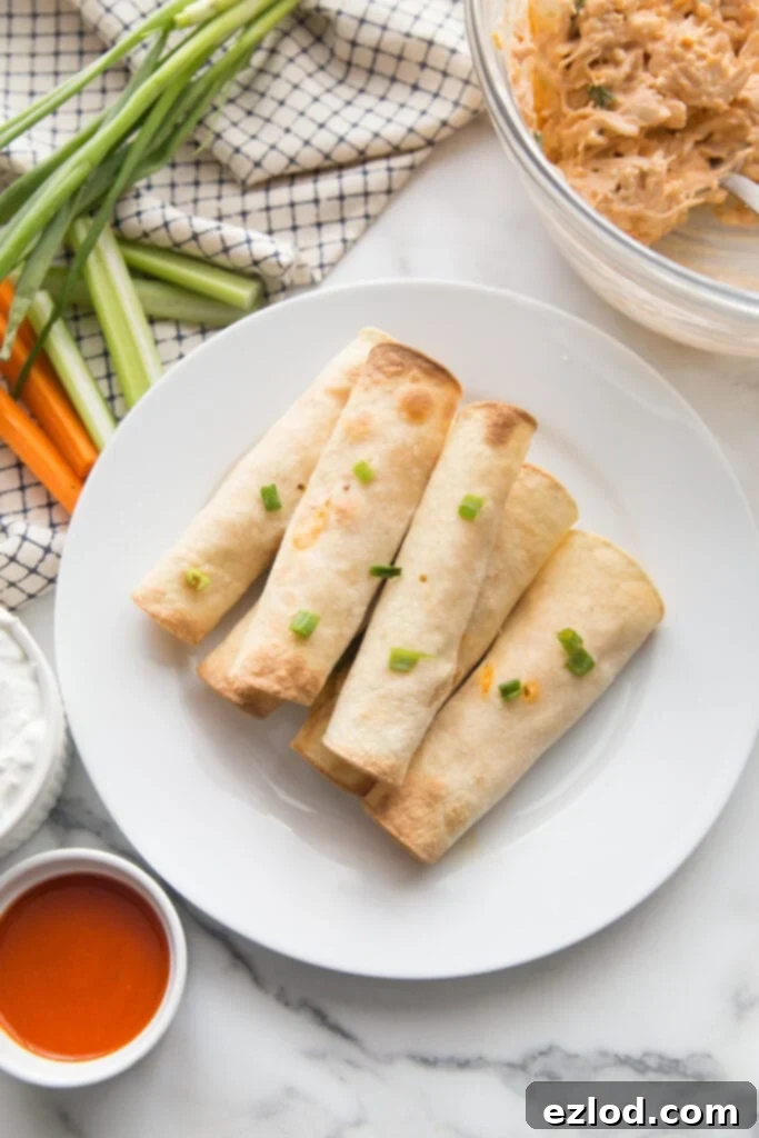 Gluten Free Buffalo Chicken Taquitos are stacked on a white dinner plate sprinkled with green onion. In the background is a glass bowl of extra buffalo chicken filling, ramekins of buffalo sauce and blue cheese dipping sauce, and celery and carrot sticks garnished with green onions..