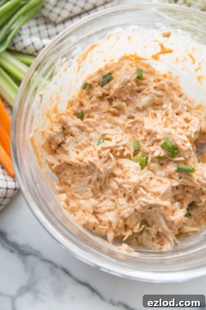 A glass bowl of buffalo chicken taquito filling topped with sliced green onions and carrots and celery sticks in the background.
