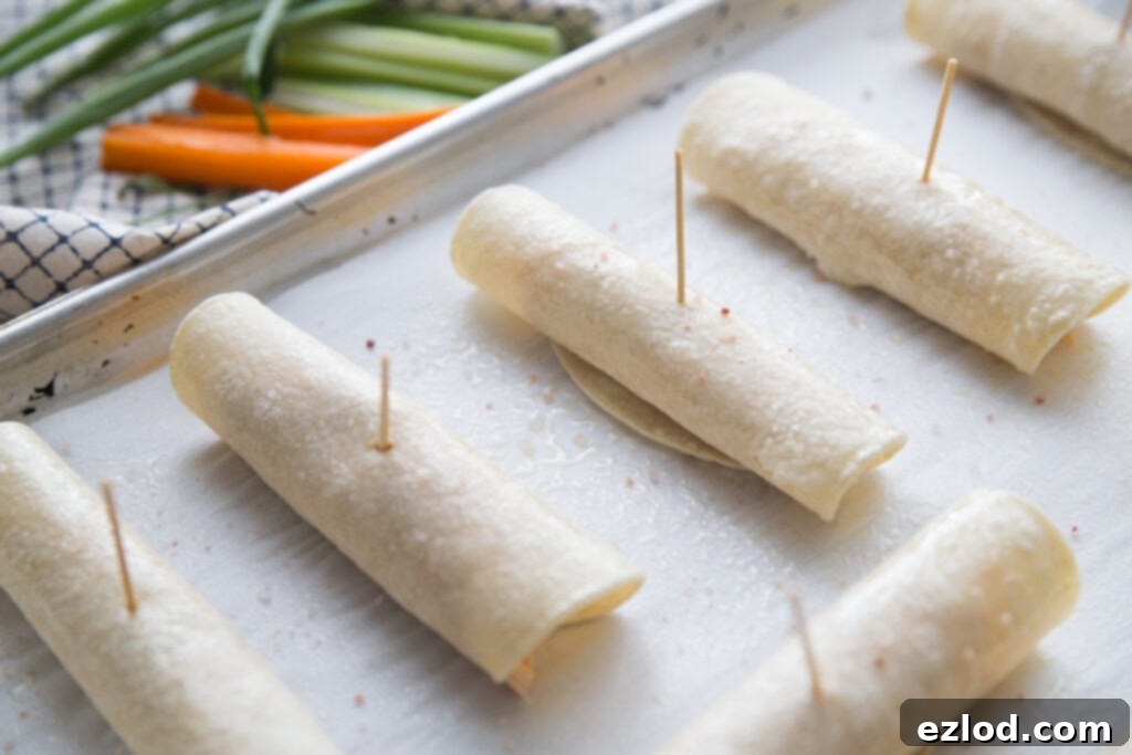 Buffalo chicken taquitos line a sheet pan ready to be put in the airfryer or oven. Each of the 6 taquitos have a toothpick securing them. In the background are celery sticks, carrot sticks, and green onions on a white and black plaid kitchen towel.