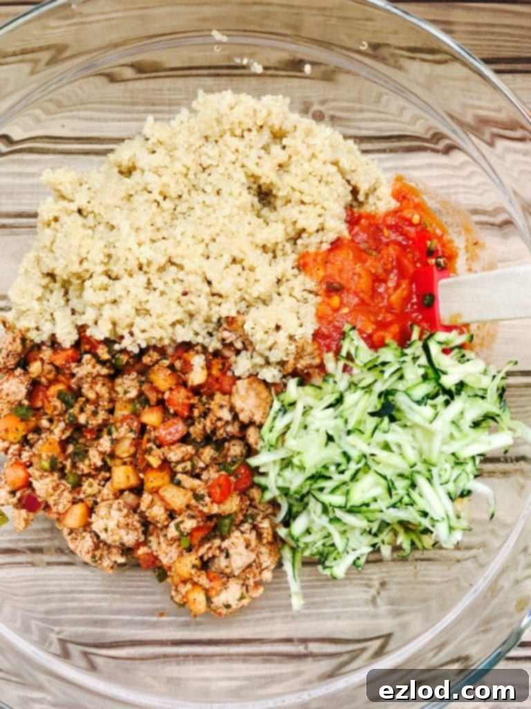 Deconstructed ingredients for a Turkey Quinoa Taco Bake- Sauteed turkey and veggies, shredded zucchini, cooked quinoa, and tomato sauce in a glass mixing bowl on a wooden surface.