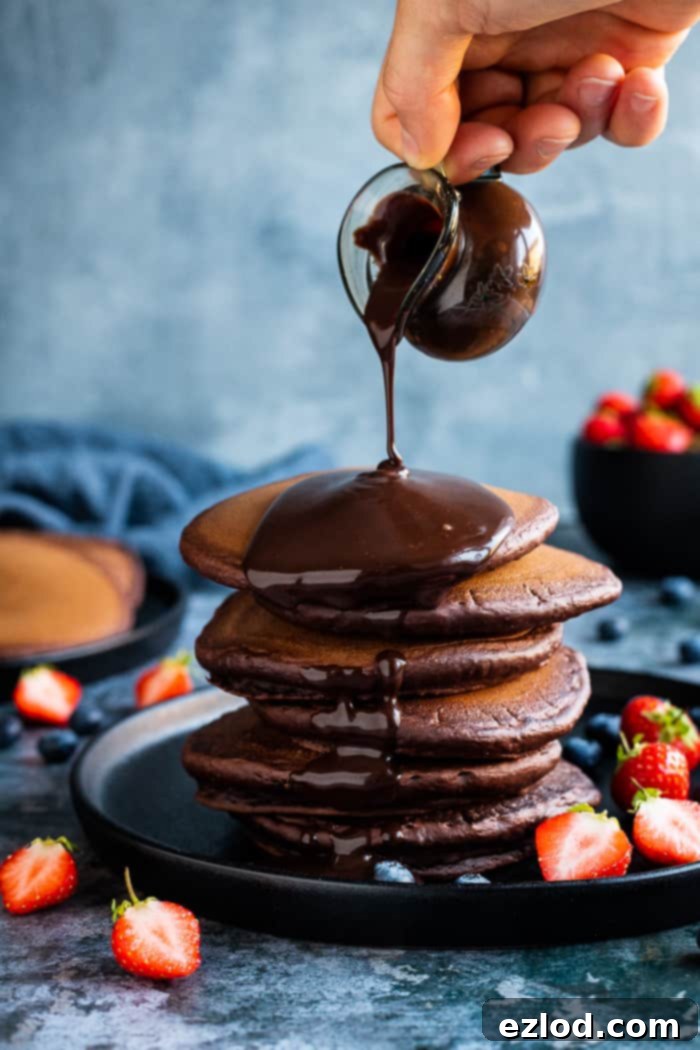 Fluffy vegan chocolate pancakes with chocolate sauce - these easy American style vegan pancakes are super soft and fluffy and very, very chocolatey! Perfect for an indulgent breakfast or brunch. Eggless and dairy-free. #veganbreakfast #veganpancakes #pancakes #breakfastrecipes #chocolate Chocolate sauce being poured onto a stack of chocolate pancakes from a small glass jug.