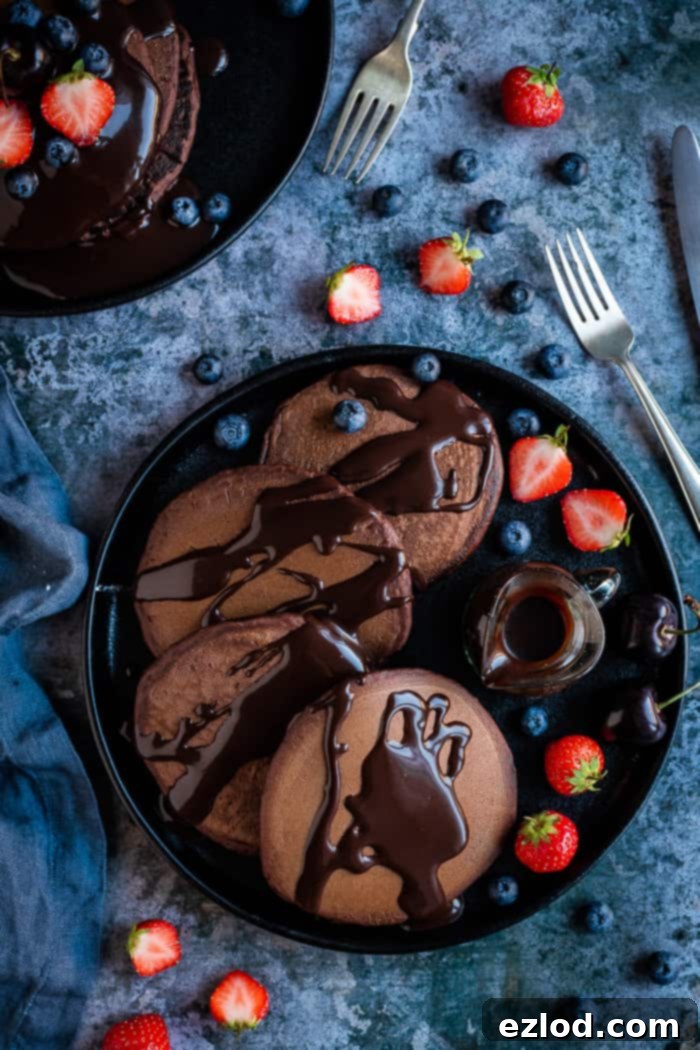 Fluffy vegan chocolate pancakes with chocolate sauce - these easy American style vegan pancakes are super soft and fluffy and very, very chocolatey! Perfect for an indulgent breakfast or brunch. Eggless and dairy-free. #veganbreakfast #veganpancakes #pancakes #breakfastrecipes #chocolate Four vegan chocolate pancakes on a black plate topped with chocolate sauce and berries on a grey background.