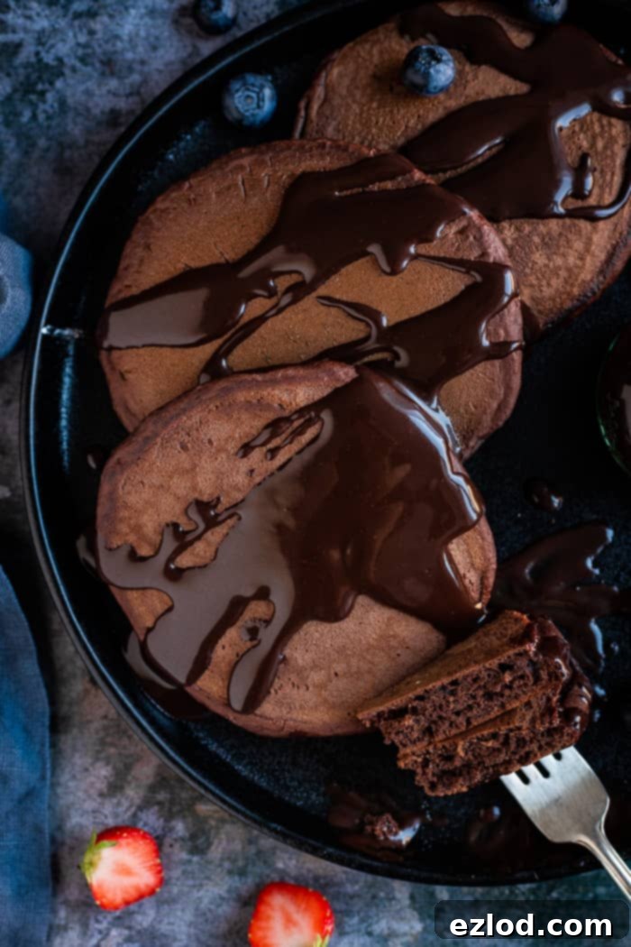Fluffy vegan chocolate pancakes with chocolate sauce - these easy American style vegan pancakes are super soft and fluffy and very, very chocolatey! Perfect for an indulgent breakfast or brunch. Eggless and dairy-free. #veganbreakfast #veganpancakes #pancakes #breakfastrecipes #chocolate Close up of chocolate pancakes with chocolate sauce on a black plate with a fork with a couple of slices of pancake on it.
