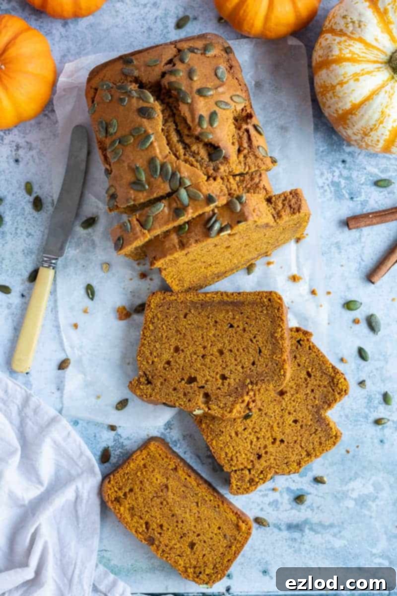 Classic Vegan Pumpkin Bread - Ideal for Fall Snacking A perfectly baked loaf of vegan pumpkin bread, generously topped with pumpkin seeds, resting on baking parchment. The background features miniature pumpkins, cinnamon sticks, a grey cloth, and a knife, evoking a cozy autumn scene.