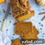 Perfect Vegan Pumpkin Bread 2 A loaf of vegan pumpkin bread, beautifully adorned with pumpkin seeds, rests on a sheet of baking parchment. The setting includes mini pumpkins, cinnamon sticks, a grey cloth, and a knife, all arranged on a blue background.
