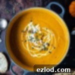 Soul-Warming Spiced Carrot and Pumpkin Soup 5 A panful of spiced carrot and pumpkin soup with a swirl of cream and pumpkin seeds on a dark background with mini pumpkins and slices of bread.