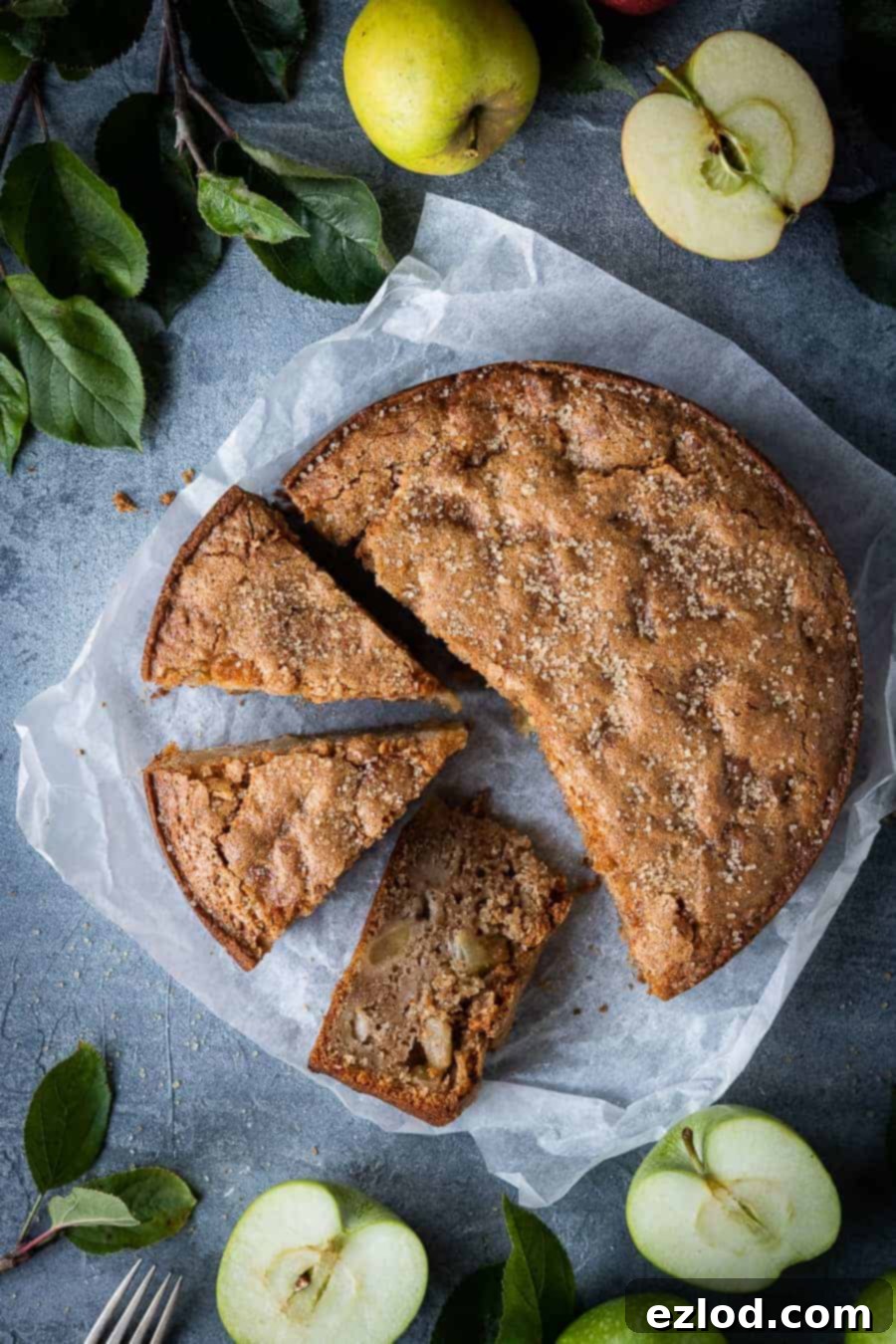 A beautifully rustic vegan apple cake, presented on a square of white baking parchment against a subtle grey background, adorned with fresh apples and vibrant apple tree leaves.