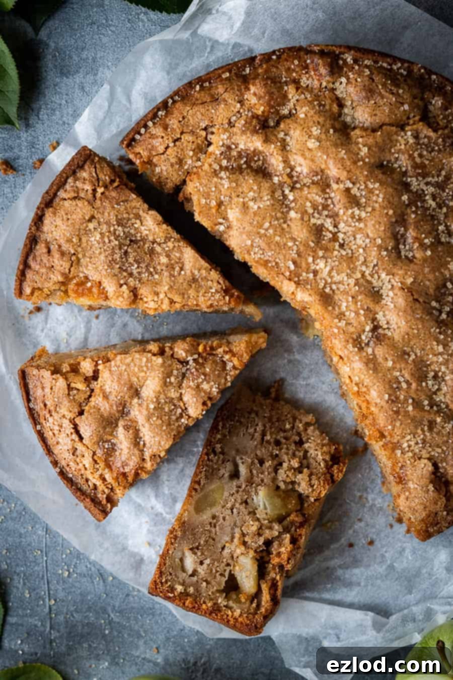 A close-up of a beautifully sliced vegan apple cake, showing the moist interior and chunks of apple, with apples and leaves in the background.