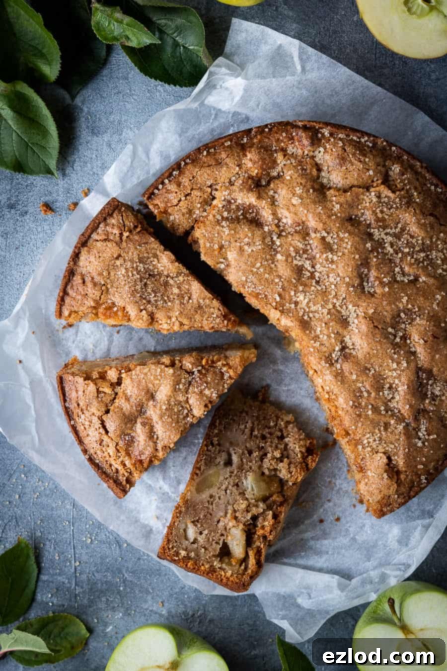 A rustic vegan apple cake, artfully arranged on a sheet of white baking parchment, surrounded by fresh apple slices and vibrant apple tree leaves.