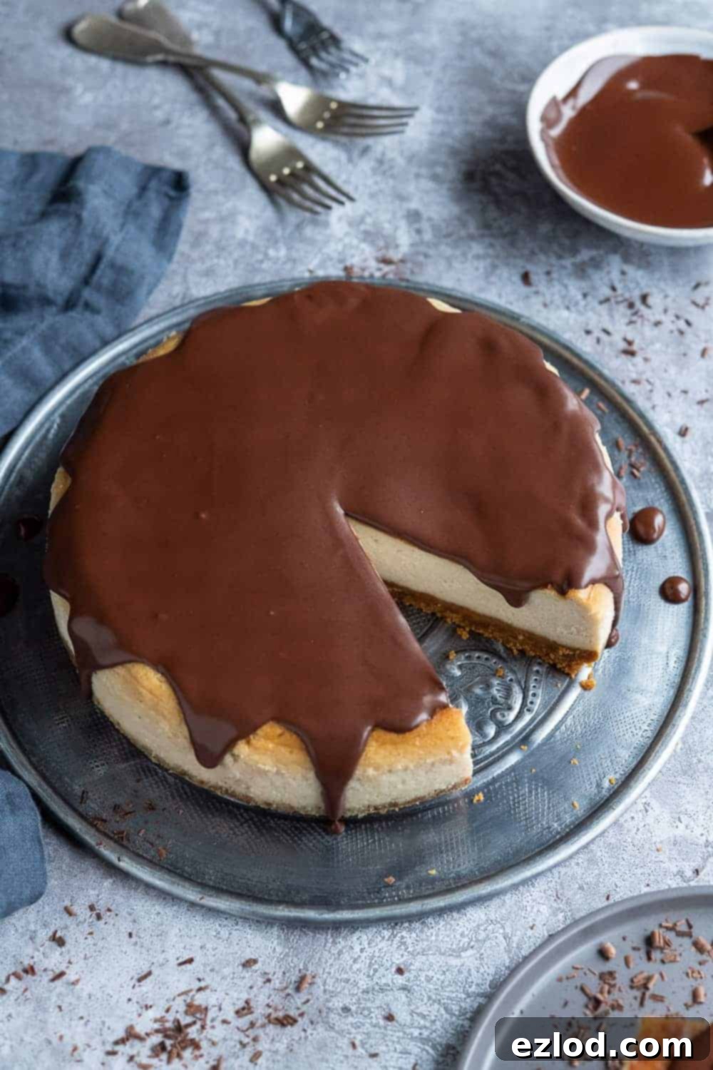 Beautifully presented Vegan Baileys Cheesecake, a perfect centerpiece dessert with its glossy ganache. A stunning vegan Baileys cheesecake, lavishly topped with chocolate ganache and chocolate shavings, presented on a round metal plate against a soft grey background, accompanied by a bowl of extra ganache and three silver forks.