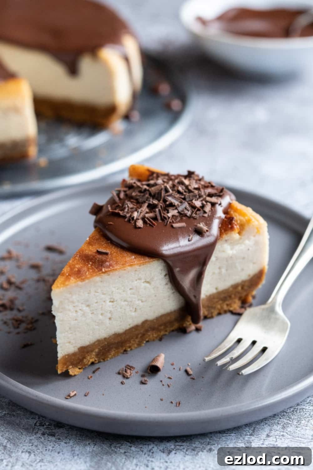 Close-up of a delightful slice of Vegan Baileys Cheesecake with ganache and chocolate shavings. A delectable slice of vegan Baileys cheesecake, elegantly garnished with a swirl of ganache and delicate chocolate shavings, served on a sophisticated grey plate with a silver fork resting beside it.