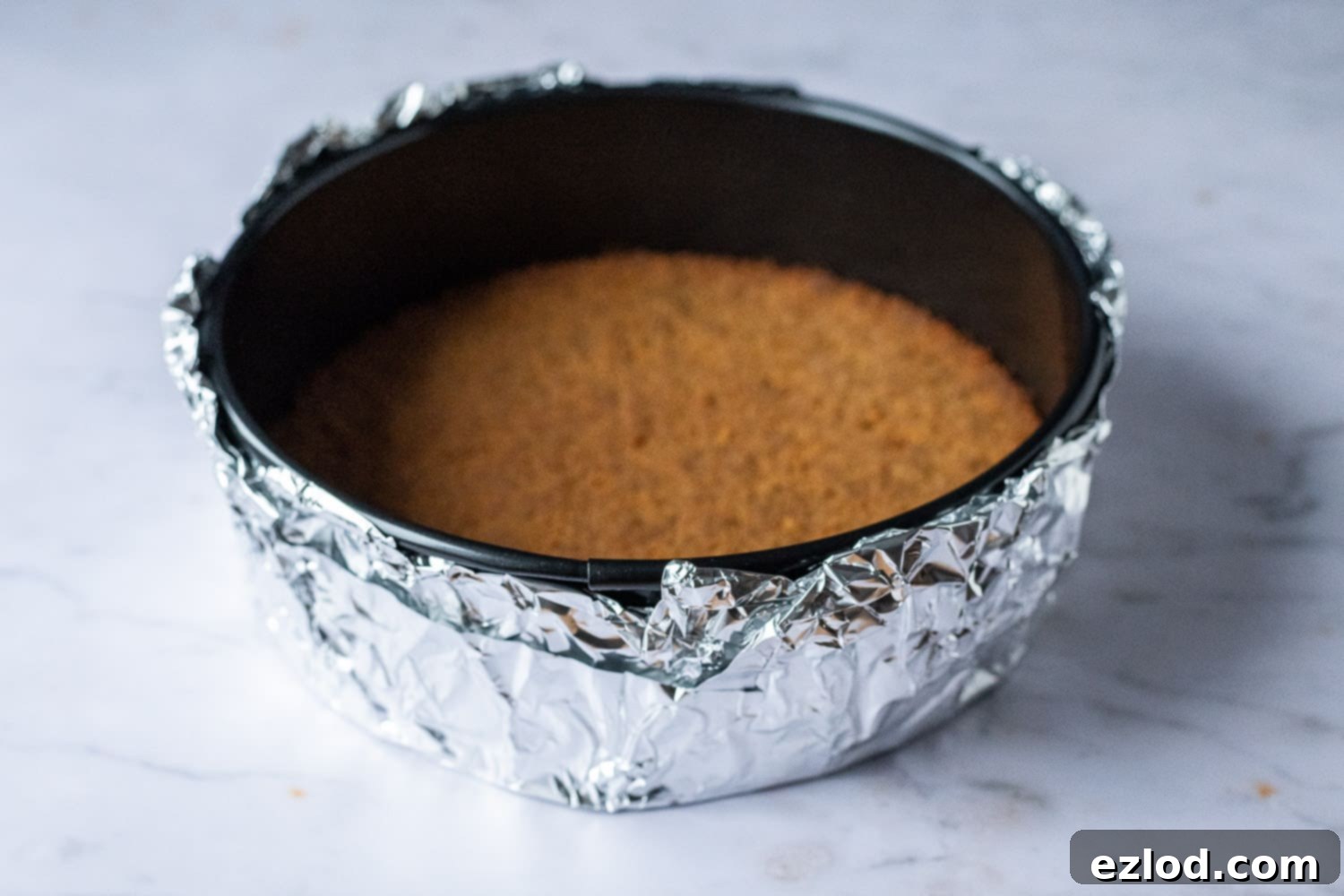 Step 3: Securely wrapping the cheesecake tin with foil for a perfect water bath bake. Step-by-step image demonstrating how to wrap the base of a springform cake tin securely with a double layer of aluminum foil for a water bath.