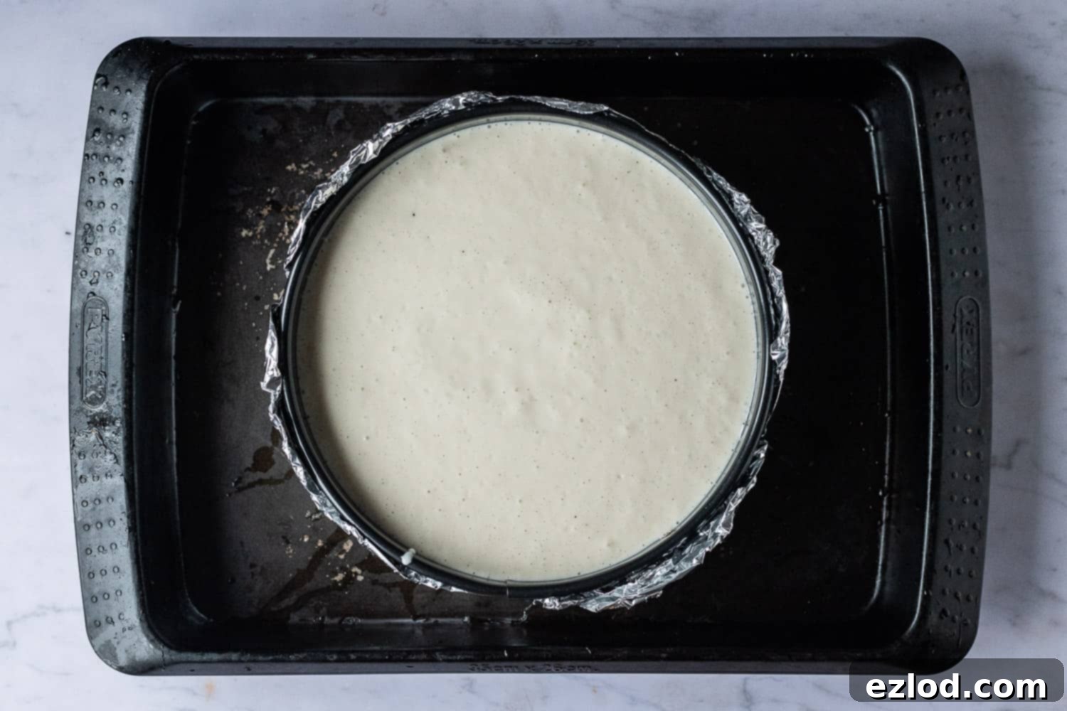 Step 4: Setting up the Vegan Baileys Cheesecake in a water bath for gentle, even baking. Step-by-step image showing the cheesecake mixture poured over the base in a foiled springform tin, placed inside a larger roasting tray, ready for the water bath.