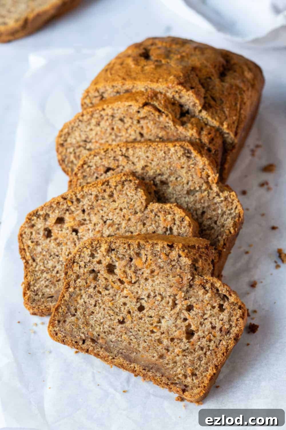 Super Moist Vegan Carrot Banana Bread - Easy Eggless and Dairy-Free Loaf Cake for Breakfast and Snacking A beautifully baked loaf of vegan carrot banana bread, perfectly sliced and arranged on a sheet of white baking parchment, showcasing its moist texture and golden-brown crust.