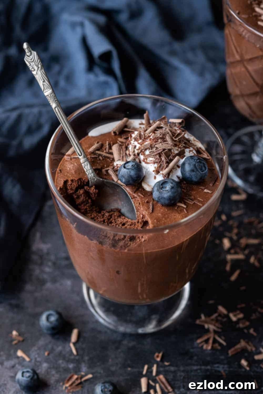 Vegan Chocolate Mousse - Light, Airy & Decadent Aquafaba Dessert A glass of vegan chocolate mousse topped with whipped cream, chocolate shavings and blueberries with a spoonful removed.