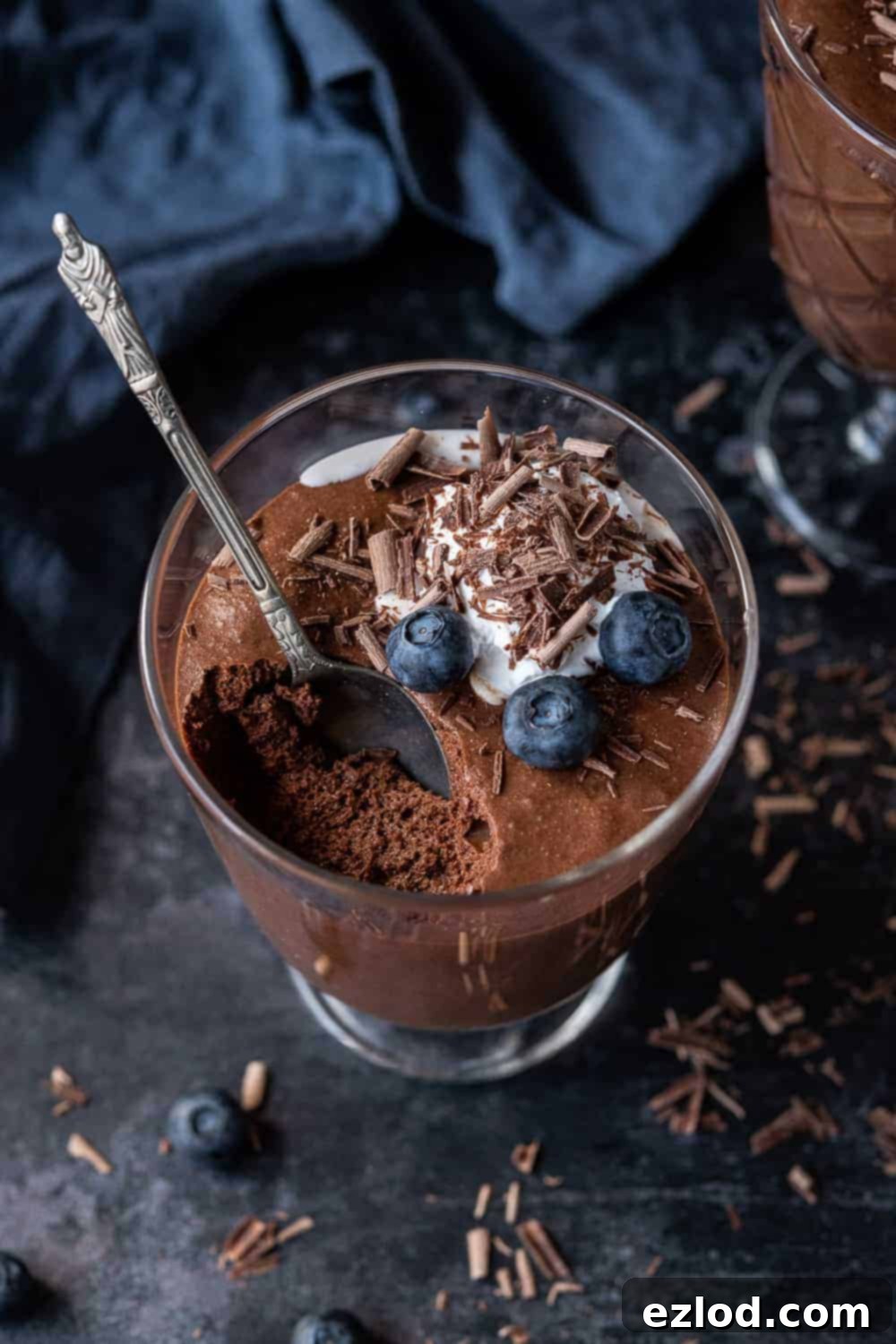 Vegan Chocolate Mousse Ready to Serve A glass of mousse on a dark background with a blue cloth and a scattering of chocolate shavings.