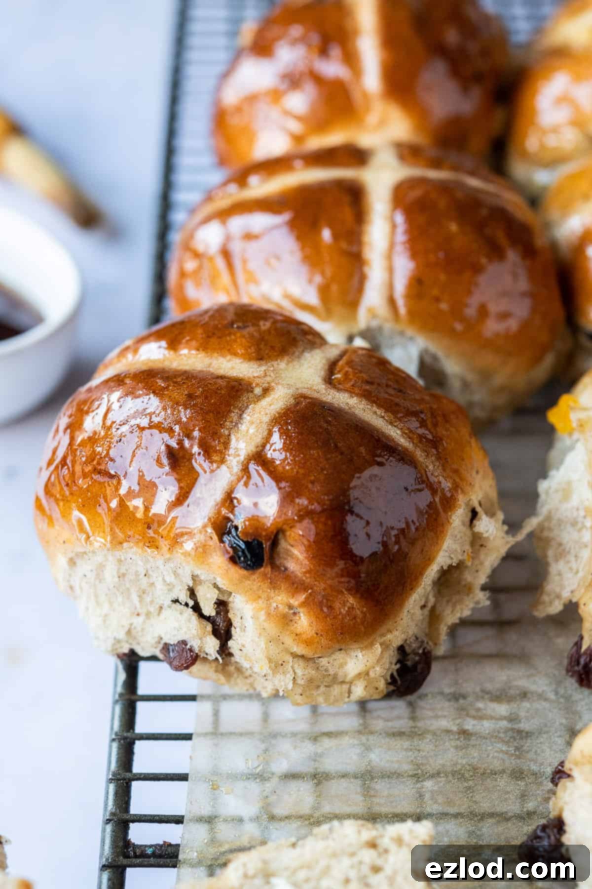 Perfectly Baked Vegan Hot Cross Bun, Glazed and Cooling. #veganbaking #easter #easterrecipes #veganbread #veganrolls Close up of a freshly baked vegan hot cross bun resting on a wire rack, showcasing its glossy finish and perfect cross. A delightful eggless and dairy-free Easter treat.