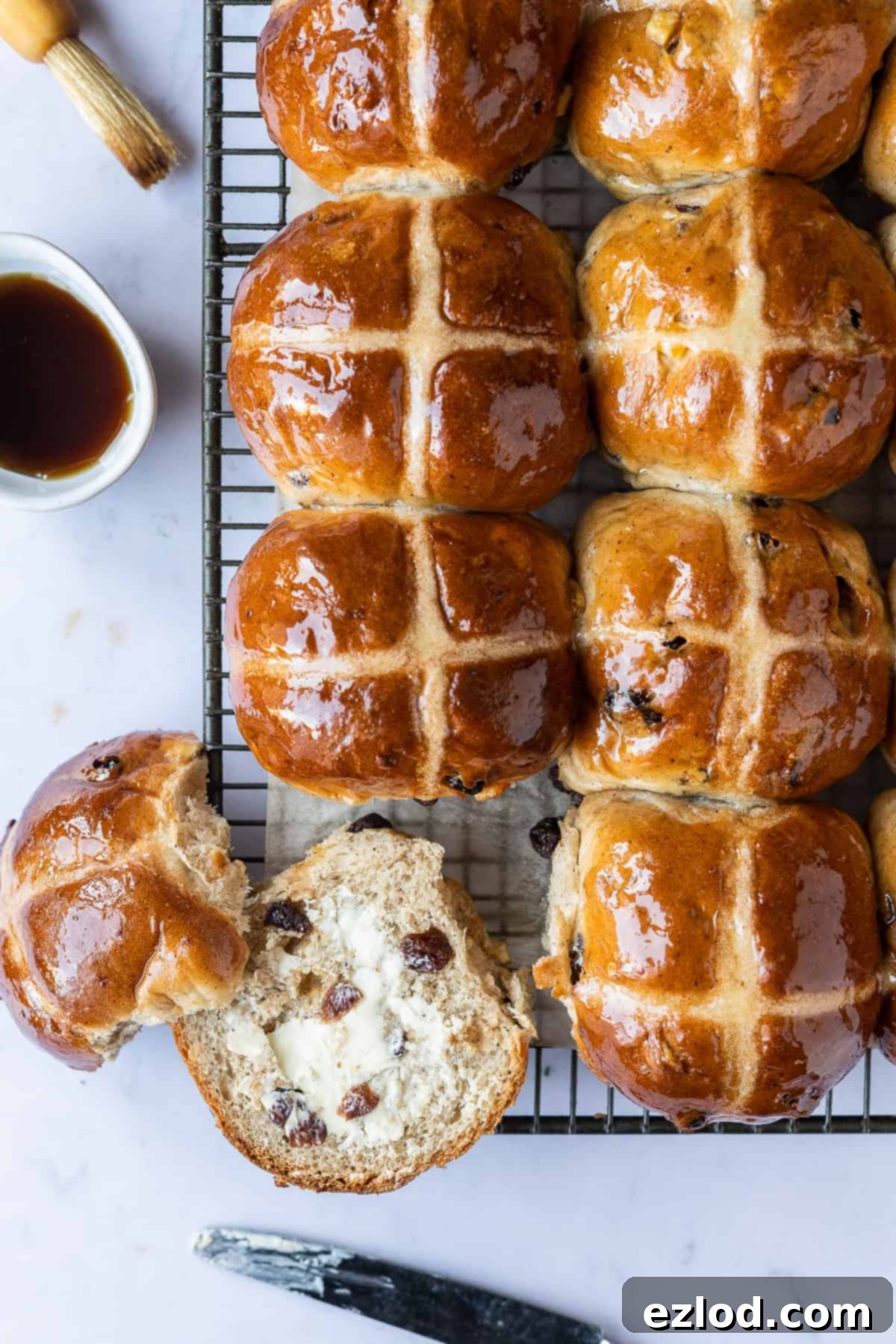 Homemade Vegan Hot Cross Buns - Eggless & Dairy-Free, Ready to Be Toasted and Enjoyed with Maple Syrup. #veganbaking #easter #easterrecipes #veganbread #veganrolls Vegan hot cross buns cooling on a wire rack, with one bun split open and buttered, alongside a small bowl of maple syrup and a pastry brush. A perfect visual for a dairy-free Easter breakfast or snack. #veganbaking #easter #easterrecipes #veganbread #veganrolls