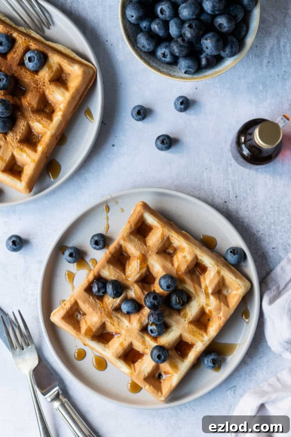 Vegan waffles - these amazing vegan waffles are crispy on the outside and soft and fluffy on the inside; the ultimate vegan breakast! Eggless and dairy free and no banana! Includes options for making them with or without aquafaba. #veganbreakfast #veganwaffles #brunch #breakfast Two golden vegan waffles presented on individual grey plates, adorned with fresh blueberries and a generous drizzle of maple syrup, ready to be enjoyed.