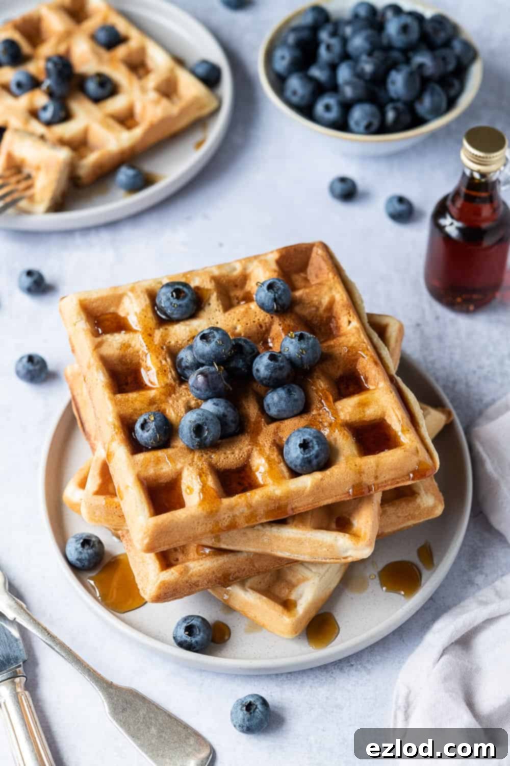 Vegan waffles - these amazing vegan waffles are crispy on the outside and soft and fluffy on the inside; the ultimate vegan breakast! Eggless and dairy free and no banana! Includes options for making them with or without aquafaba. #veganbreakfast #veganwaffles #brunch #breakfast A beautifully arranged stack of golden vegan waffles on a grey plate, adorned with a scattering of fresh blueberries and a generous drizzle of rich maple syrup, ready to be enjoyed as a perfect vegan breakfast or brunch.