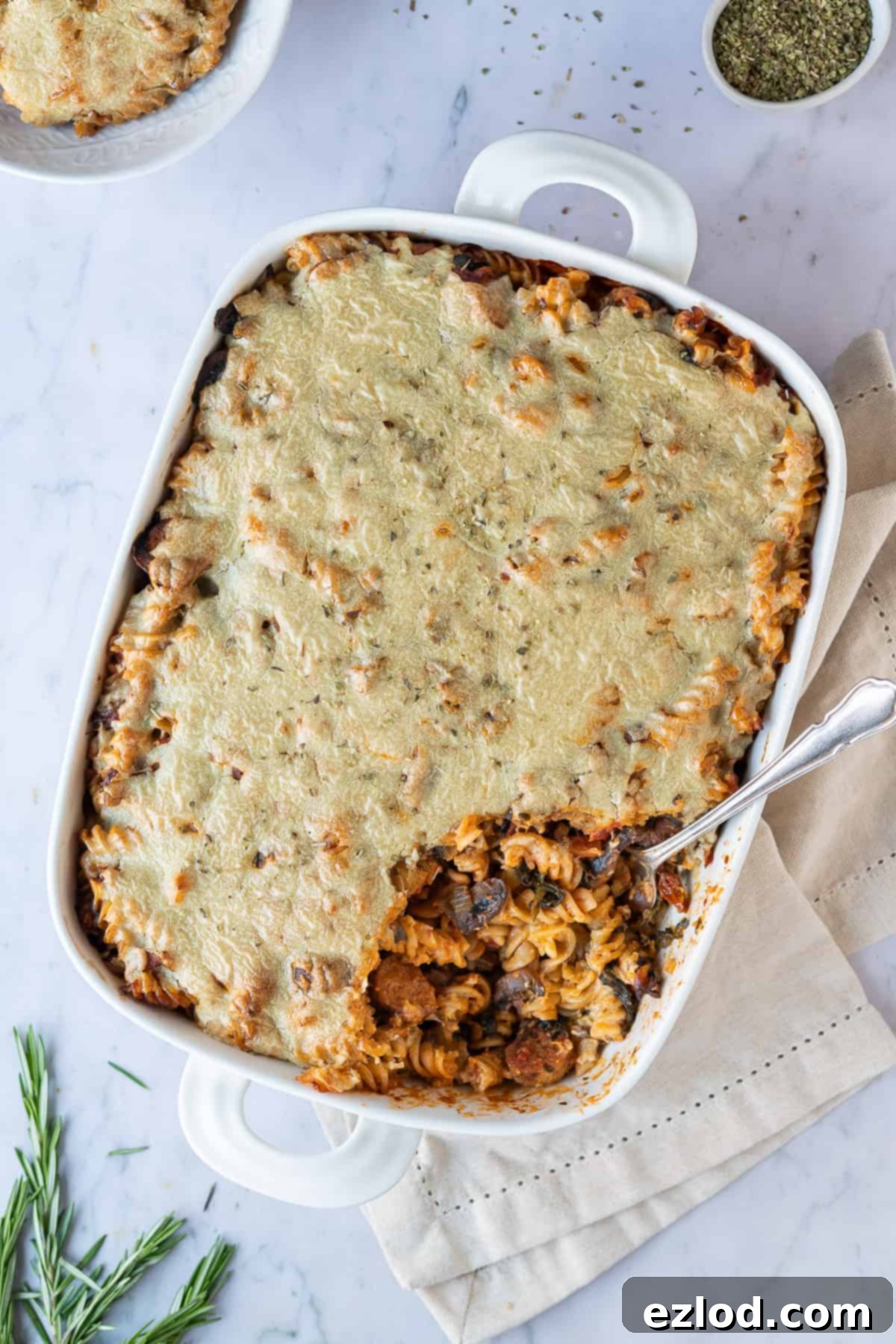 Vegan Pasta Bake with Cashew Cheese A large white baking dish with vegan pasta bake, set on a marble background, garnished with fresh rosemary and a small bowl of oregano.