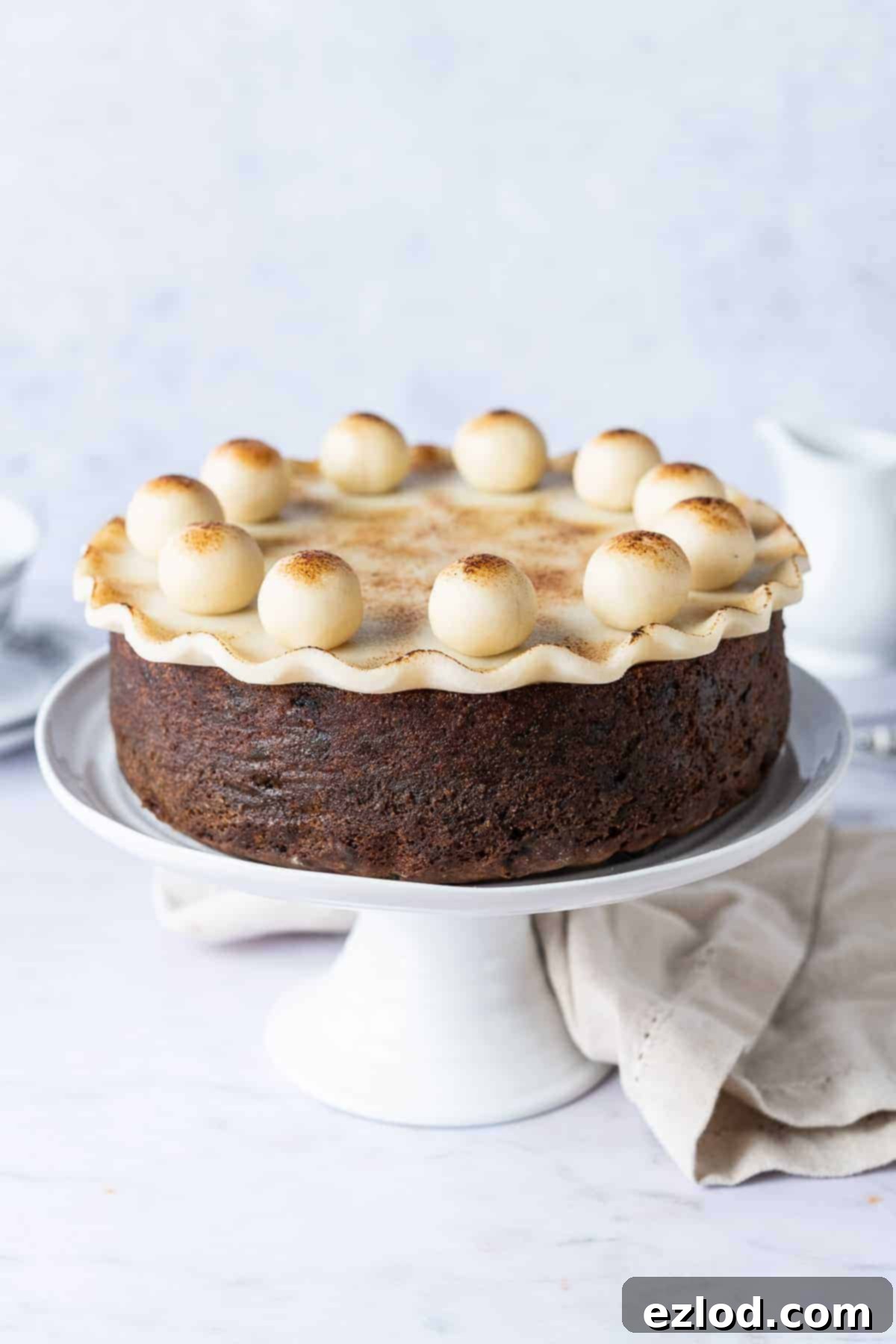 Vegan Simnel Cake: Eggless & Dairy-Free British Easter Fruit Cake with Marzipan Decorations A beautifully decorated vegan Simnel cake, showcasing its golden marzipan topping and eleven marzipan balls, displayed on a white cake stand against a soft grey background, accompanied by a cream cloth, a white milk jug, and teacups, evoking a cozy Easter tea scene.