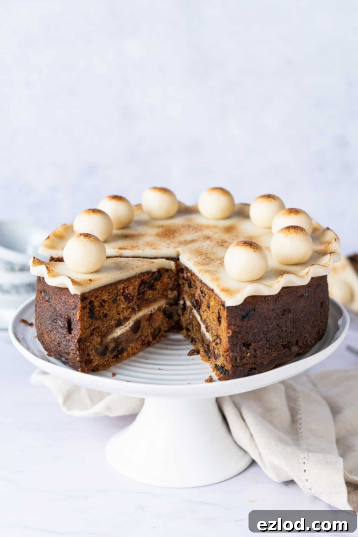 Whole Vegan Simnel Cake Ready for Easter Celebration A magnificent whole vegan Simnel cake, freshly baked and elegantly decorated, resting on a pristine white cake stand against a soft, light grey background, highlighting its festive charm and readiness for celebration.