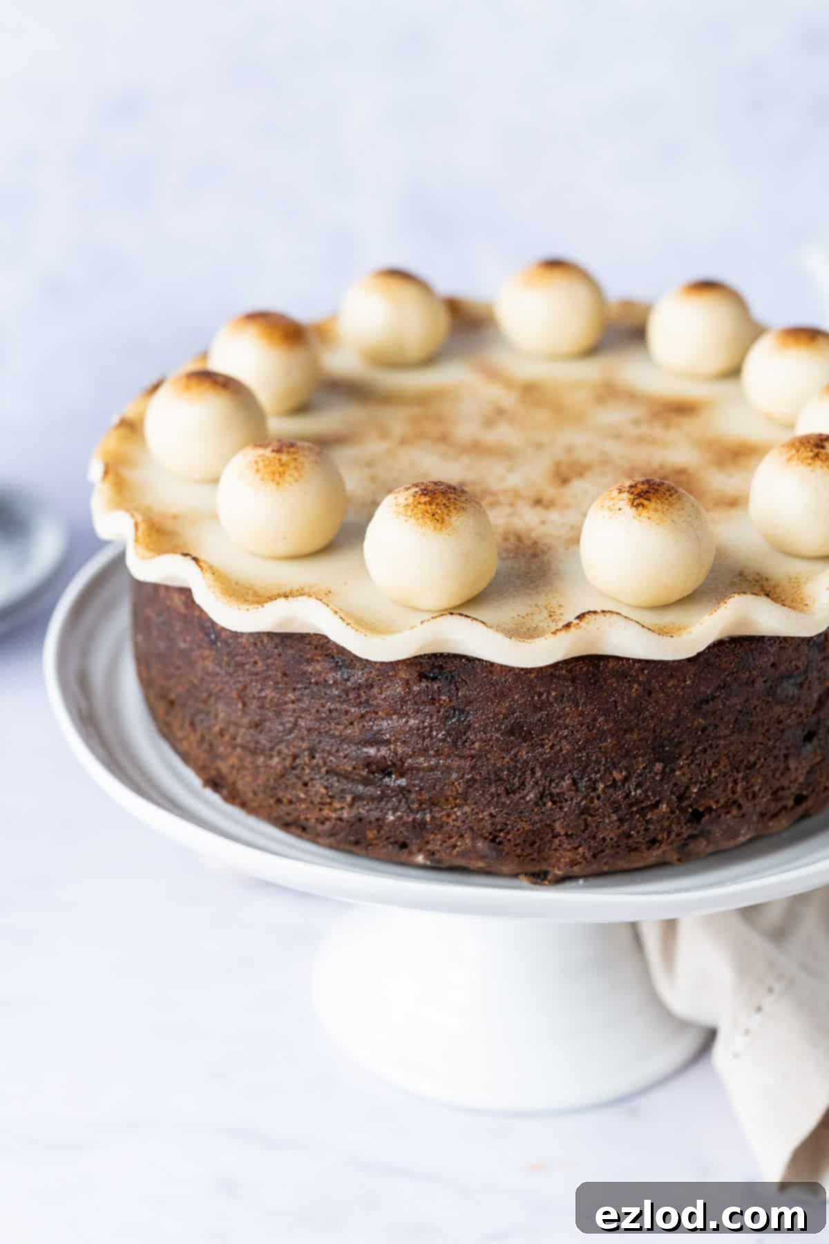Decorative Vegan Simnel Cake with Toasted Marzipan Topping A beautiful close-up of a fully decorated vegan Simnel cake, highlighting the golden-brown toasted marzipan topping and the traditional eleven marzipan balls arranged in a perfect circle, resting on a white cake stand.